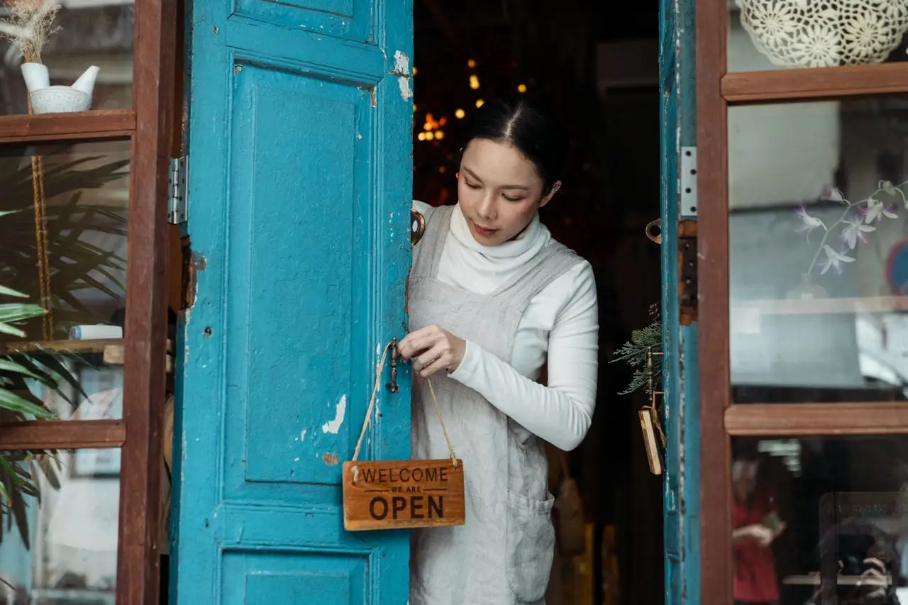 A woman adjusts an open sign on a blue wooden door of a cafe inviting customers inside