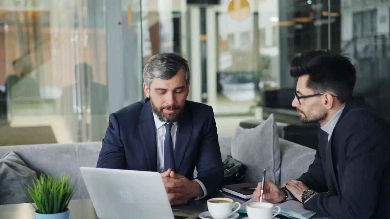 Two businessmen in suits discussing work at a cafe with laptops and coffee